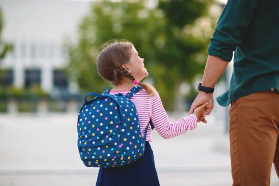 Ein kleines Mädchen mit einem gepunkteten Rucksack hält die Hand eines Erwachsenen, während sie gemeinsam im Freien spazieren, symbolisch für das Umgangsrecht und die Betreuung von Kindern.